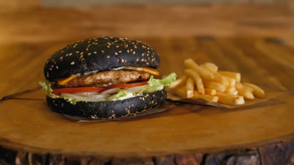 Handen in handschoenen pick-up een sappige zwarte hamburger met schnitzels en groenten. Ongezonde fastfood. Franse frietjes blijven op het houten bord. — Stockvideo