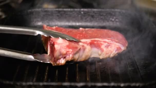 Un trozo de carne cruda gira por pinzas metálicas en una sartén caliente. Cocinar T-Bon Steak de cerca. Deliciosa comida chatarra grasa . — Vídeos de Stock