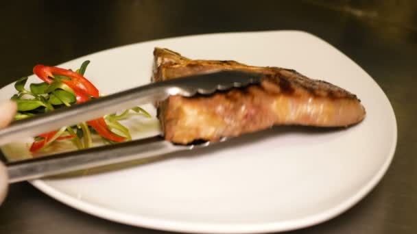 Свежеприготовленный стейк из t-bone выложен на белой тарелке рядом с кучей микрозелени. Подготовка готового куска мяса к подаче. Вкусная жирная вредная пища — стоковое видео