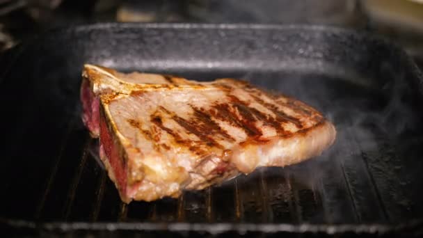 Um pedaço de carne e vire um osso por pinças de metal em uma panela de grelha quente. Cozinhar T-Bon Steak Closeup. Deliciosa comida gordurosa — Vídeo de Stock