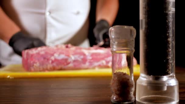 Um cozinheiro em um avental branco limpa um pedaço de carne crua de gordura usando faca. Em primeiro plano, há sal e pimenta nos moinhos. Açougueiro vai fazer carne de porco . — Vídeo de Stock