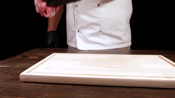 Ένας μάγειρας με λευκή ποδιά βάζει ένα κομμάτι ωμό κρέας σε μια ξύλινη σανίδα, αλάτι, εποχή και να το πάρει στο πλάι. — Αρχείο Βίντεο