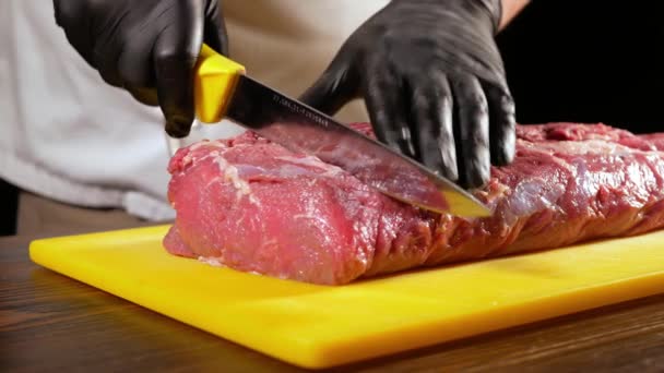 En professionell slaktare i svarta handskar skivor en bit rå kött för biff från Entrecote med en vass kniv. Kocken gör en förberedelse för matlagning på grillen. — Stockvideo