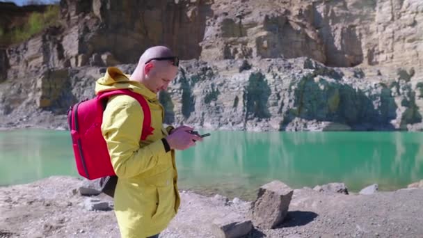 Unge man styr en drönare med hjälp av en fjärrkontroll med joysticks. Drönarpiloten letar efter en andrepilot på himlen. En snygg kille med en röd ryggsäck klädd i en gul regnrock står emot — Stockvideo