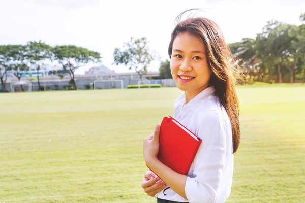 Portrait of young student smiling holding notebook at outdoor under sunlight on greenfield in the university