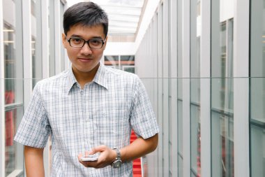 Yakışıklı Asyalı çocuk gülümsüyor ve koridordaki akıllı telefonunu kontrol ediyor.
