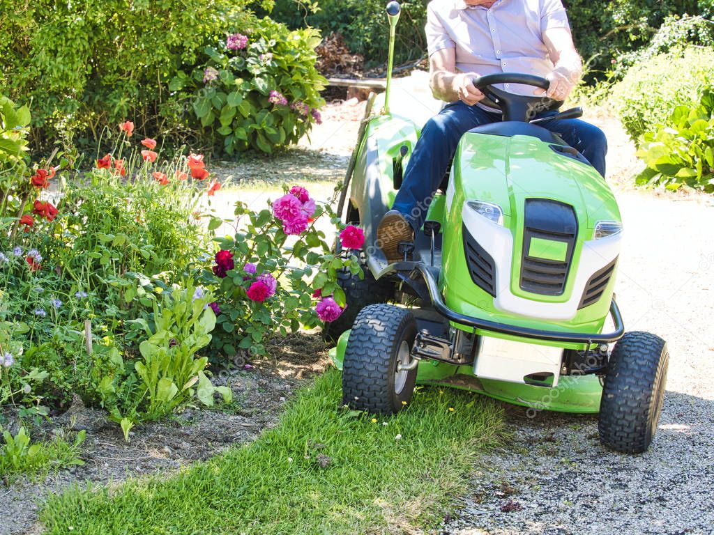 Senior man driving a tractor lawn mower in garden with flowers