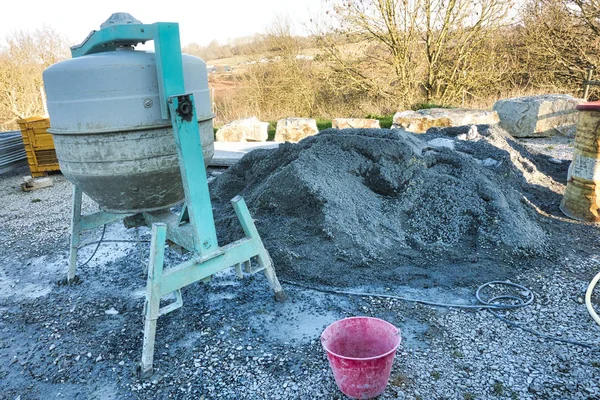 Cement machine close to bunch of gravel with red bucket of water.