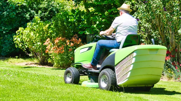75歳の老人は 花のある庭でトラクター芝刈り機を運転しています 緑と白のパワーライド カラフルな花の間のフィールドに回転 — ストック写真