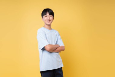 Parlak bir gülümsemesi olan genç bir çocuk sarı arka planda kollarını kavuşturarak güvenle ayakta duruyor.