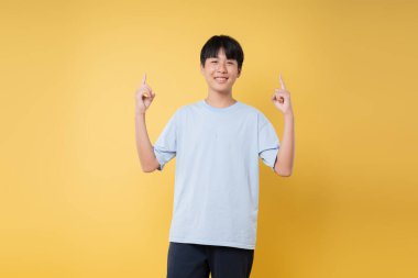 Gülümseyen bir genç iki eliyle işaret ediyor. Sarı bir arka planda gelişigüzel giyinmiş.