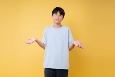 Genç bir çocuk, sorgulayıcı bir ifadeyle dimdik ayakta duruyor ve sarı bir zeminin önünde omuzlarını silkiyor.