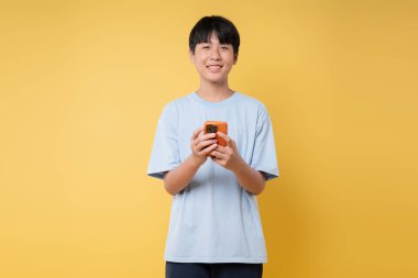 Mavi tişörtlü, gülümseyen genç, telefonunu stüdyoda, parlak sarı bir arka planda tutuyor.