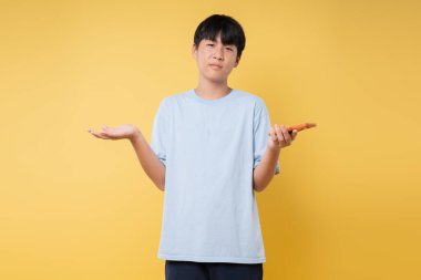 Çocuk sarı arka planda turuncu bir nesne tutuyor. Bir kolu havada ve açık mavi tişört giymiş yüzü sorguluyor.