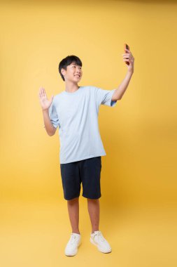 Tişört ve şort giyen, selfie çeken, gülümseyen ve sarı bir fonun önünde el sallayan genç bir çocuk.