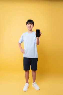 Genç bir Asyalı çocuk, bir stüdyo çekiminde koyu sarı bir arkaplanda siyah ekran akıllı telefon gösteriyor.