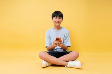 Sarı bir zeminde bağdaş kurmuş oturan genç Asyalı bir çocuk elinde telefonla içten bir şekilde gülümsüyor.