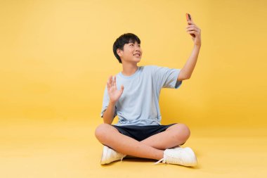 Bir video görüşmesi sırasında parlak bir arka planda günlük giysiler giyerek bacak bacak bacak üstüne atarak gülümseyen bir çocuk.