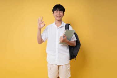 Sırt çantası ve kitapla gülümseyen öğrenci, sarı arka plana karşı hazırlıklı olduğunu belirten iyi bir jest yaptı.