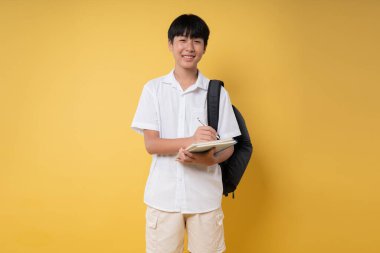 Sırt çantası taşıyan neşeli genç bir öğrenci ve sarı bir arka plana dizilmiş bir defter.