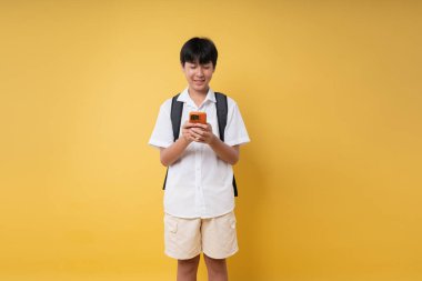 Beyaz gömlekli ve sırt çantalı bir öğrenci canlı sarı bir arkaplan önünde telefonlarını kontrol ediyor.