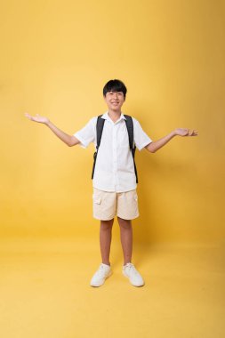 Sırt çantalı üniformalı bir öğrenci mutlu bir şekilde sarı bir arka planda okula hazır bekliyor.