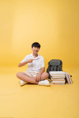 Gülümseyen öğrenci, okul çantasının yanındaki kitapları işaret ederek sarı bir arkaplanda okumaya hazır bir öğrenciyi işaret ediyor.