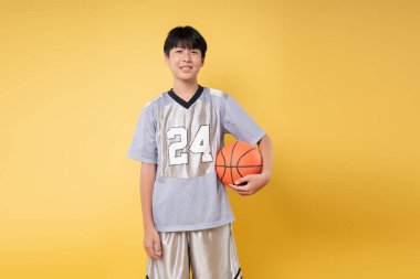 Spor giyimli genç, elinde turuncu bir basketbol topuyla saf sarıya karşı gülümsüyor.