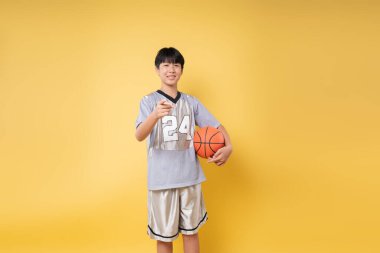 Stüdyoda basketbol topu tutan basketbol formalı bir çocuk sarı bir arka fona karşı gülümsüyor.