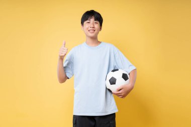 Günlük kıyafetlerle gülümseyen genç bir çocuk bir futbol topu tutuyor ve sarı bir arka plana başparmak işareti veriyor.