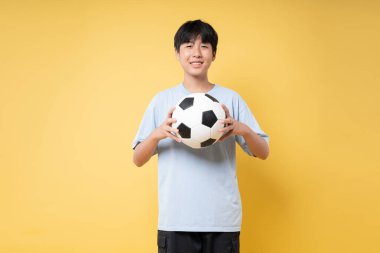 Asyalı bir genç, futbol topunu stüdyoda parlak sarı bir zemine tutarken gülümsüyor.