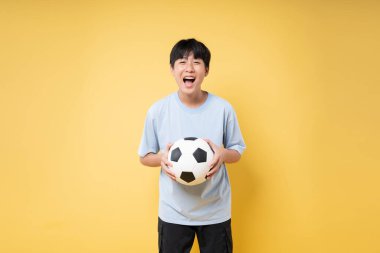 Mavi tişörtlü gülümseyen çocuk elinde futbol topuyla sarı arka planda duruyor.