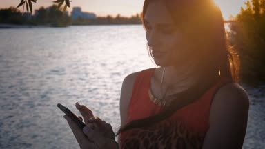 Çekici genç kadın smartphone günbatımında kullanır. Sevimli bir kız gülümsüyor ve bir cep telefonu deniz arka plan kullanır.