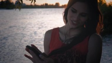 Çekici genç kadın smartphone günbatımında kullanır. Sevimli bir kız gülümsüyor ve bir cep telefonu deniz arka plan kullanır.