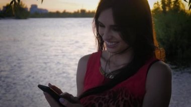 Çekici genç kadın smartphone günbatımında kullanır. Sevimli bir kız gülümsüyor ve bir cep telefonu deniz arka plan kullanır.