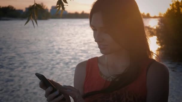 Jovem atraente usa smartphone ao pôr do sol. Uma menina bonita sorri e usa um celular no fundo do mar . — Vídeo de Stock