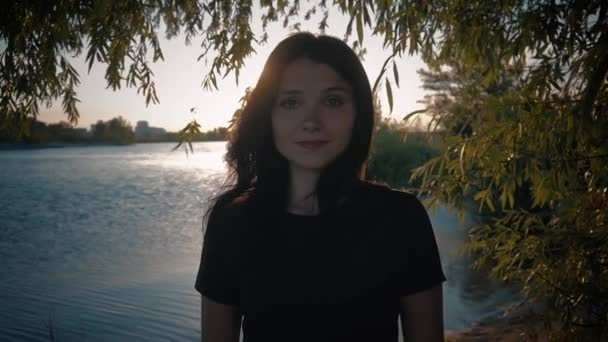 Retrato de una joven guapa morena sobre un fondo al atardecer en un lago — Vídeo de stock
