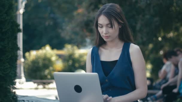 Jonge vrouw die werkt met de laptop in een park op een bankje op de straat achtergrond van een stad — Stockvideo