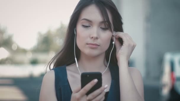 Porträtt av ung vacker kvinna med smartphone och lyssna på musik i hörlurar utomhus på gatan bakgrund — Stockvideo