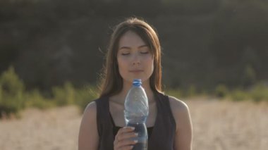 Genç kadın gezgin içme suyu plastik bir şişe sıcak kumlu alan, doğada. Kız duygu susuzluk ile su molasına gün batımında