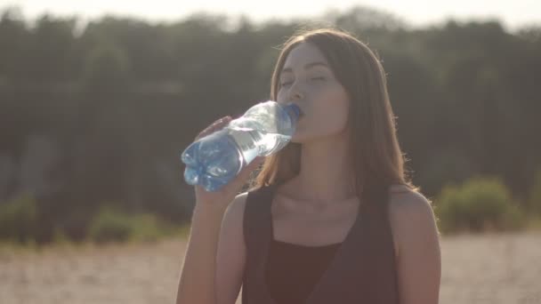 Jovem mulher que está com sede em um dia quente de verão bebe água de uma garrafa de plástico no fundo do pôr do sol . — Vídeo de Stock