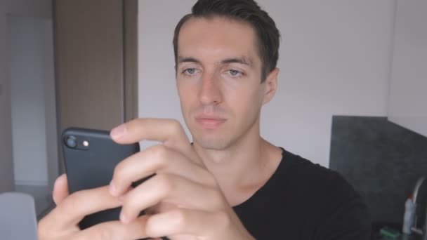 Молодий чоловік у чорній футболці використовує смартфон вдома на кухні . — стокове відео