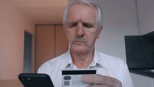 年配の男性が台所で自宅でスマート フォンを使用してクレジット カードでオンライン ショッピングします。オンラインバン キングの携帯アプリ — ストック動画