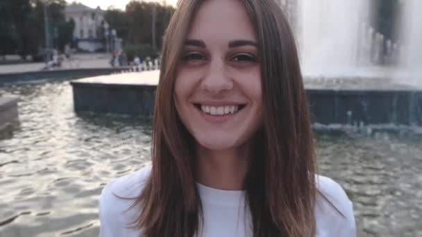 Schöne, glückliche Frau mit braunen Augen, die auf dem Hintergrund eines Stadtbrunnens auf der Straße lächelt. fröhliches Mädchen lacht Flirts — Stockvideo