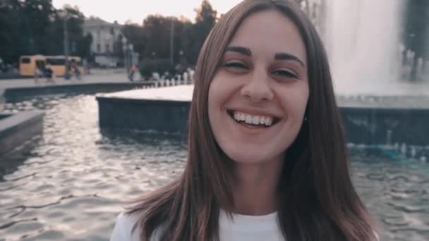 Beautiful happy woman with brown eyes smiling on a background of a city fountain in the street. Cheerful girl laughs flirts — Stock Video
