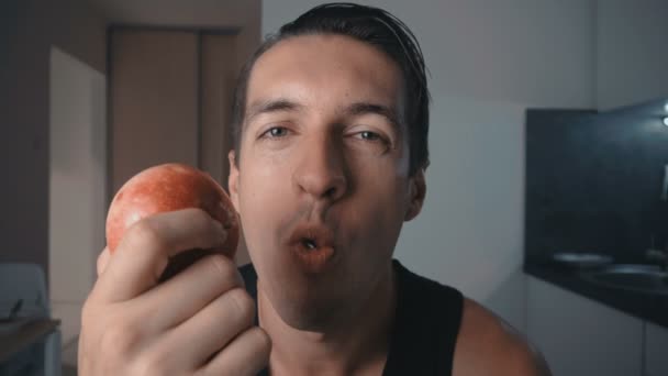 Jovem comendo maçã vermelha em casa na cozinha. Retrato de cara comendo uma maçã fresca e olhando para a câmera . — Vídeo de Stock