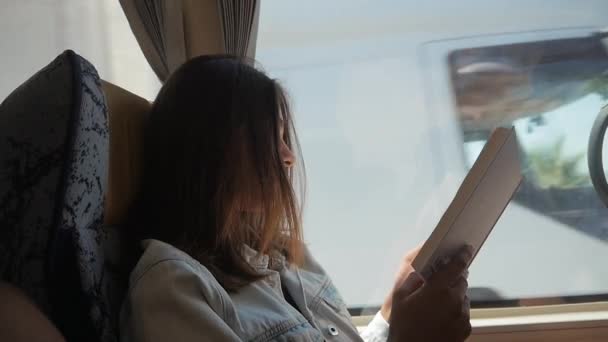 Молодая женщина путешествует на автобусе и читает книги. Девушка едет в машине перед окном — стоковое видео