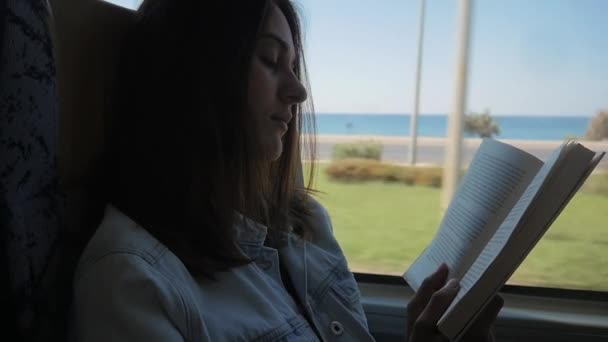Mujer joven viajando en autobús y leyendo libro. Chica está viajando en el coche delante de la ventana — Vídeo de stock