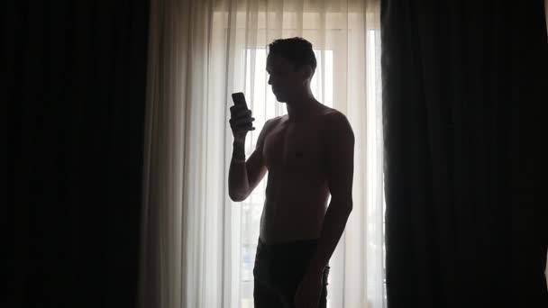 Junger Mann textet zu Hause am Fenster auf Smartphone — Stockvideo