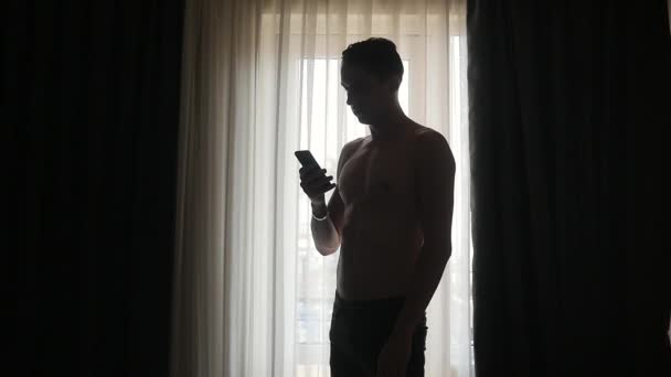 Hombre joven mensajes de texto en el teléfono inteligente por ventana en casa — Vídeos de Stock